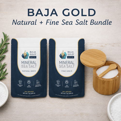 Two bags of BAJA GOLD Mineral Sea Salt, one Natural Grain and one Fine Grain, displayed on a light wood background alongside a bamboo salt container, a wooden spoon, and bowls of salt.