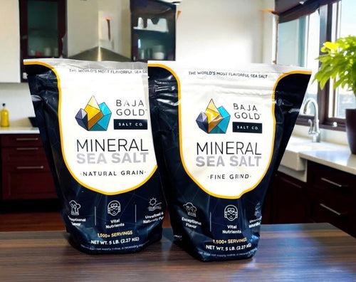 Two large bags of Baja Gold Salt Co. Mineral Sea Salt, Fine Grind, standing on a dark wooden countertop in a kitchen setting.