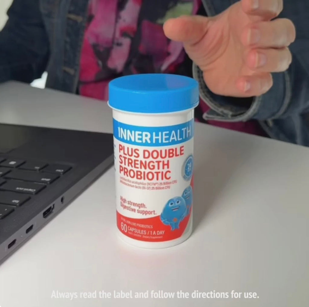 A person seated at a desk holds a bottle of INNER HEALTH Plus Double Strength Probiotic. The bottle has a blue lid and a white label with red and blue text and graphics.